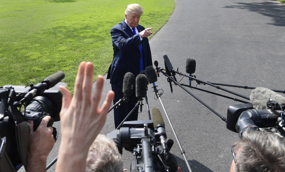 Donald Trump stands outside pointing while a group of media members stand in the foreground with microphones and cameras.