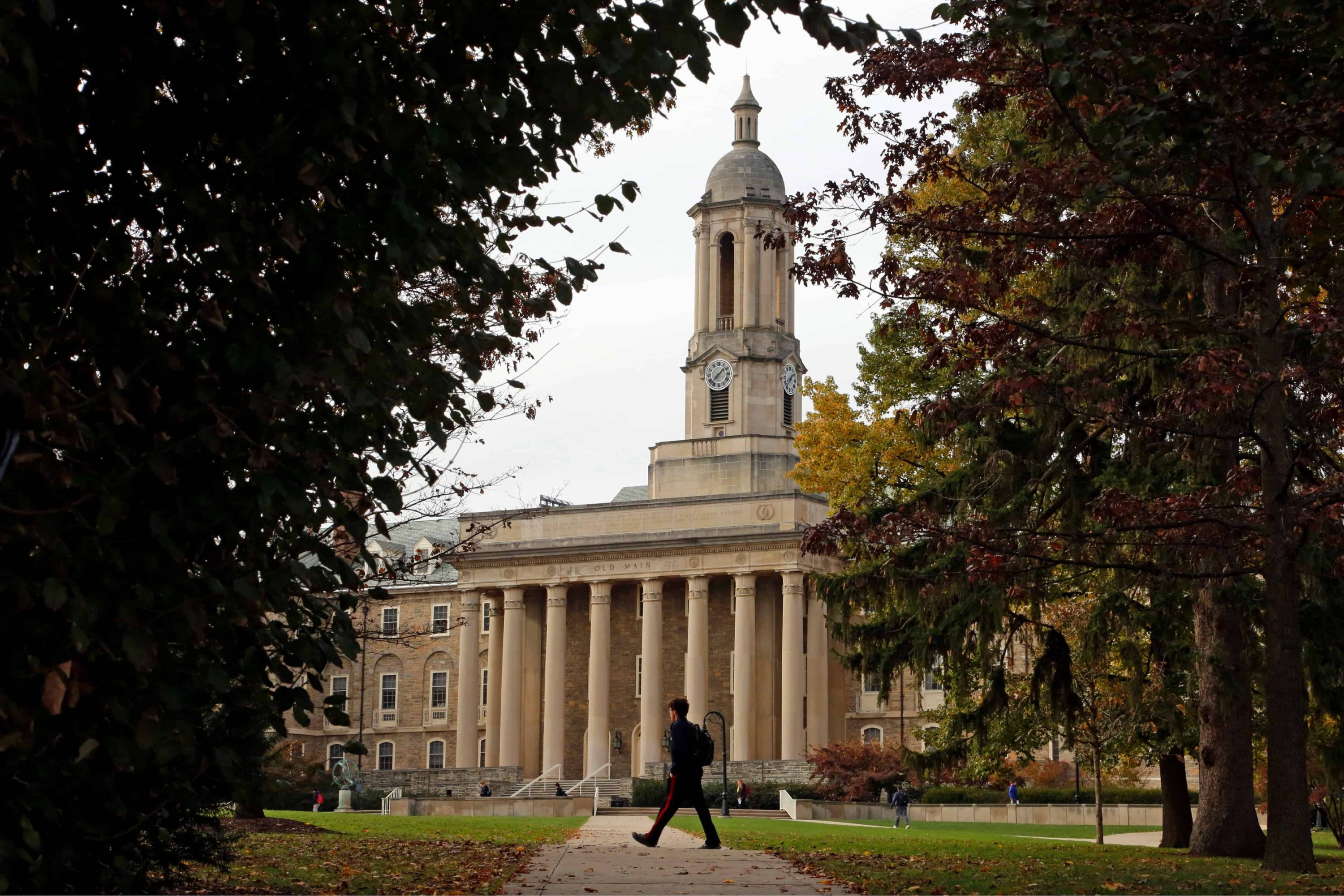 A student walks on a grassy quad at Penn State University with a classical building with a clock tower in the distance.