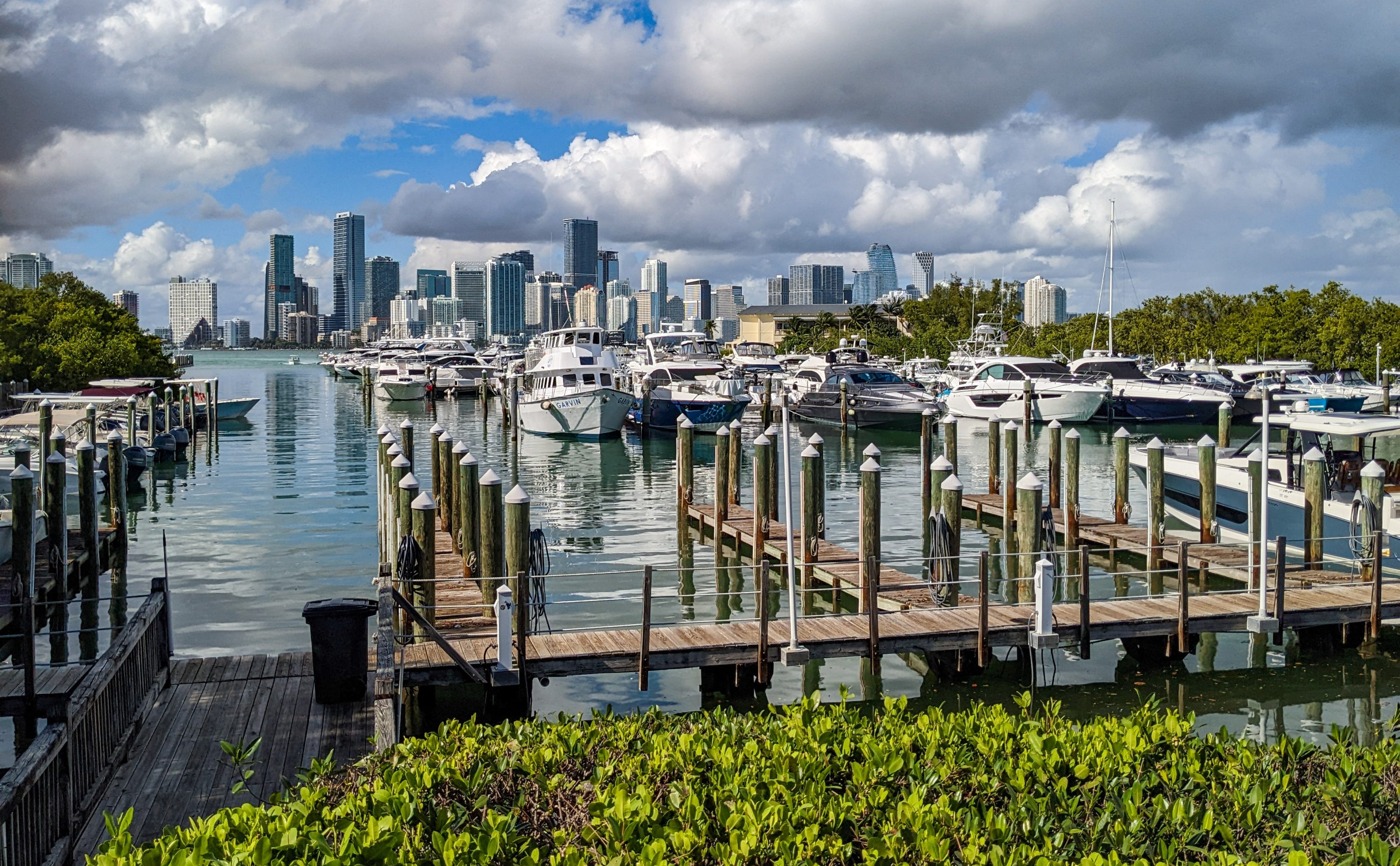 Several boats float in water by docks in front skyscrapers in the background.